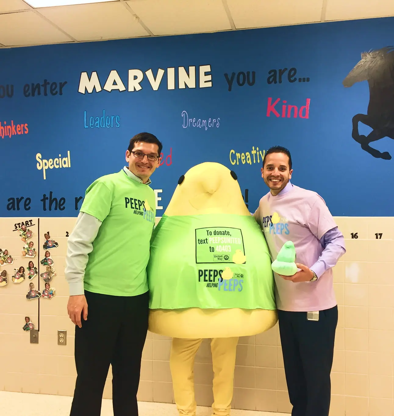 Two men and a large yellow Peeps marshmallow mascot stand in front of a blue mural at Marvine.