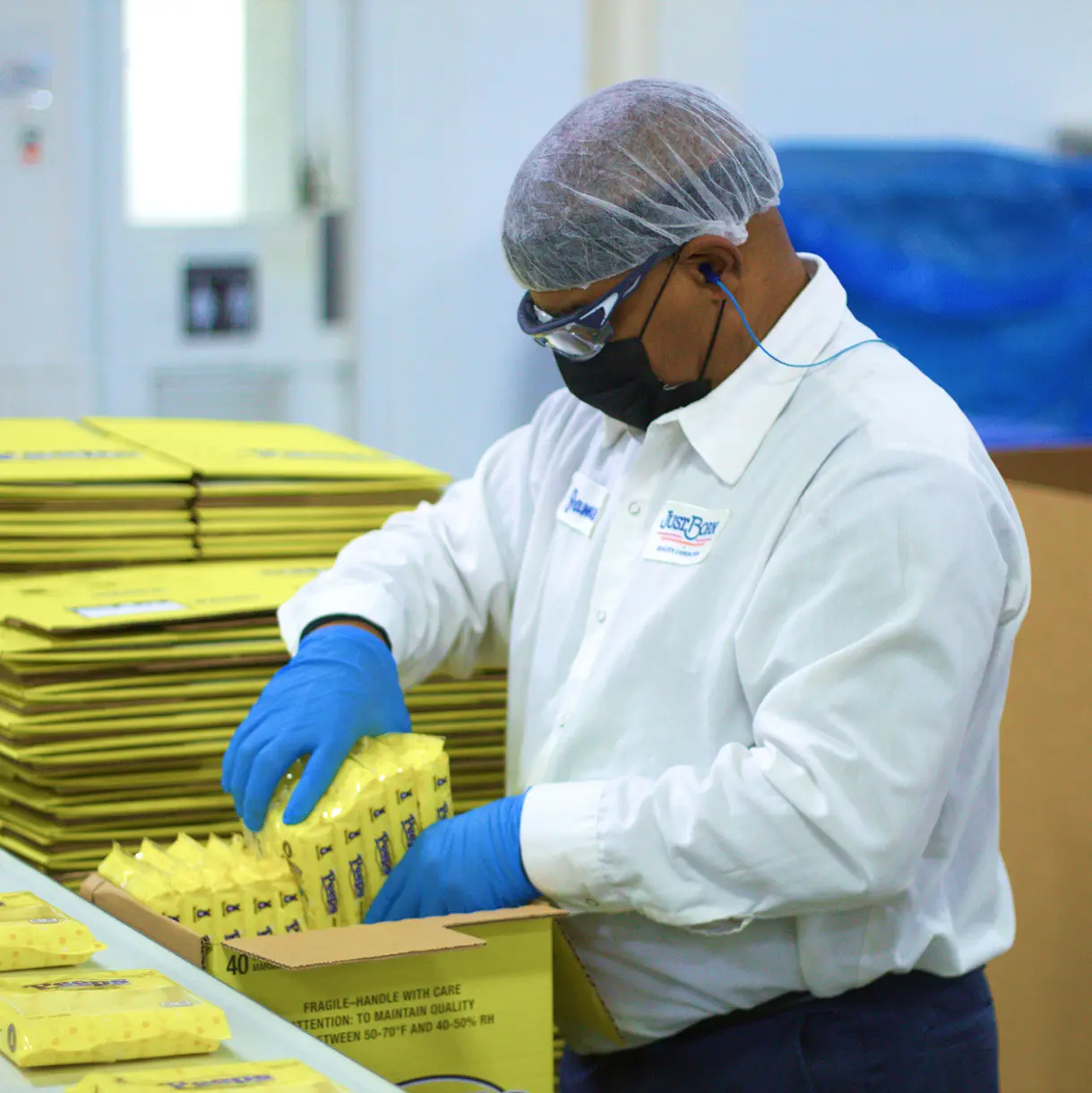 A worker in a hairnet, mask, and blue gloves packs yellow products into a box in a clean manufacturing facility.