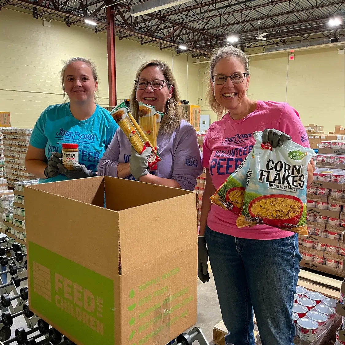 Three volunteers smiling while packing food at a warehouse. They hold pasta, corn flakes, and canned goods next to a "Feed the Children" box.