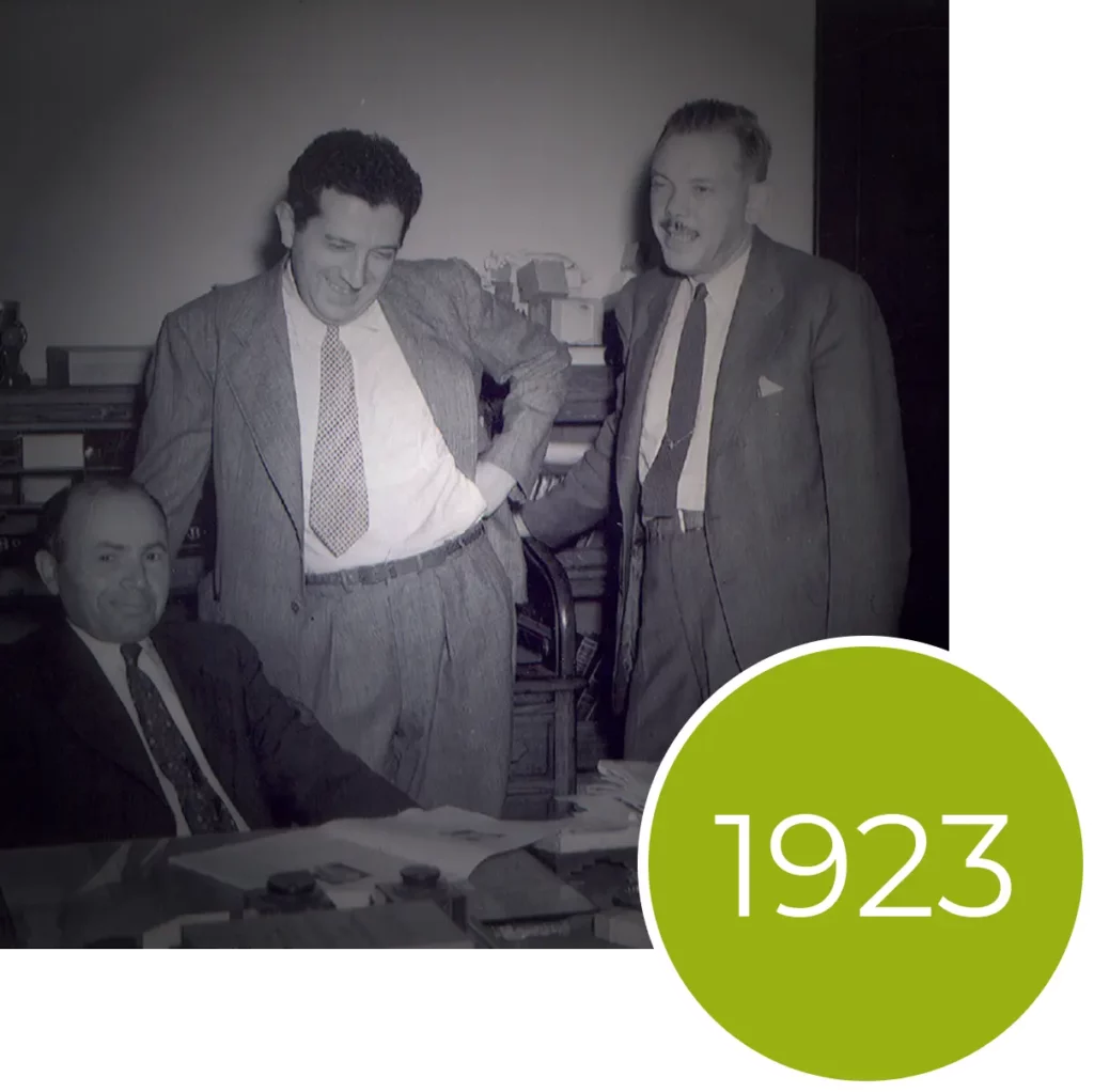 Three men in suits gather around a desk in an office with a green circle overlay reading 1923.