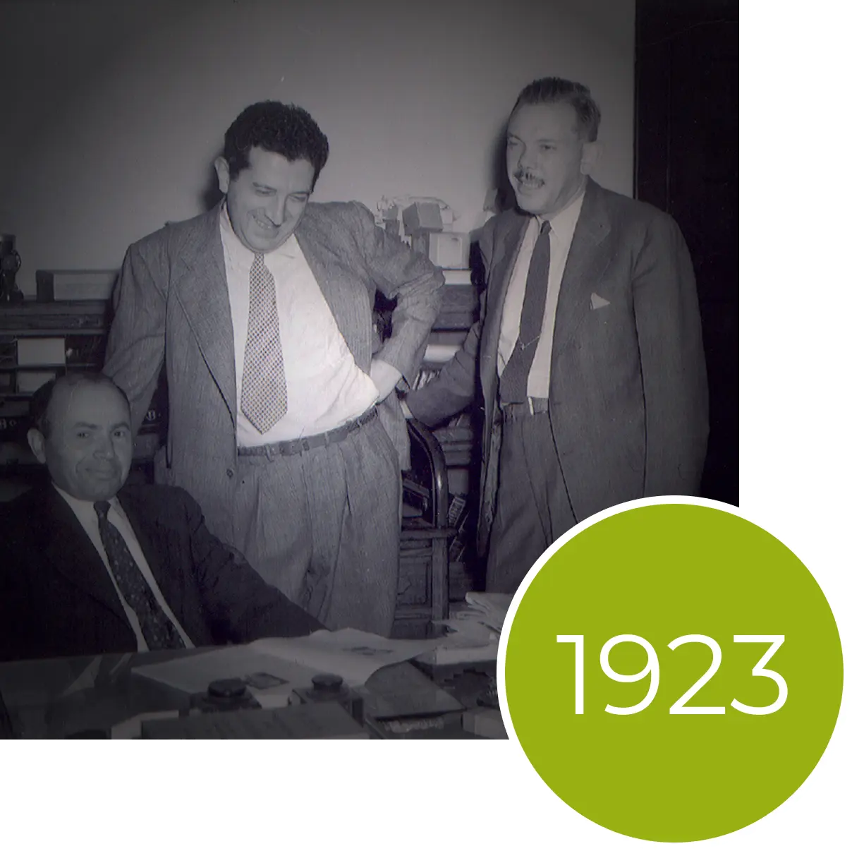 Three men in suits gather around a desk in an office with a green circle overlay reading 1923.