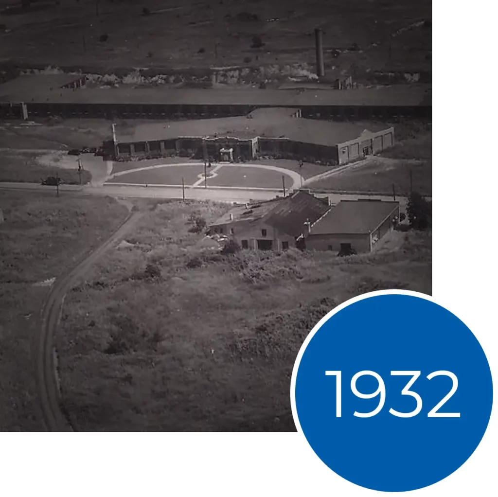 Aerial black and white photo of a 1932 industrial building complex with a circular driveway and blue text overlay.