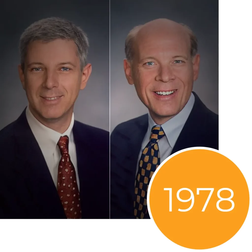 Two side-by-side portraits of smiling men in suits with a large orange circle containing the year 1978.