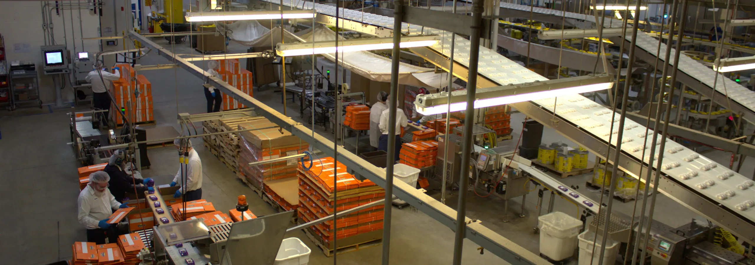 Workers in a large packaging facility handling orange boxes on an assembly line with complex conveyor belt systems.
