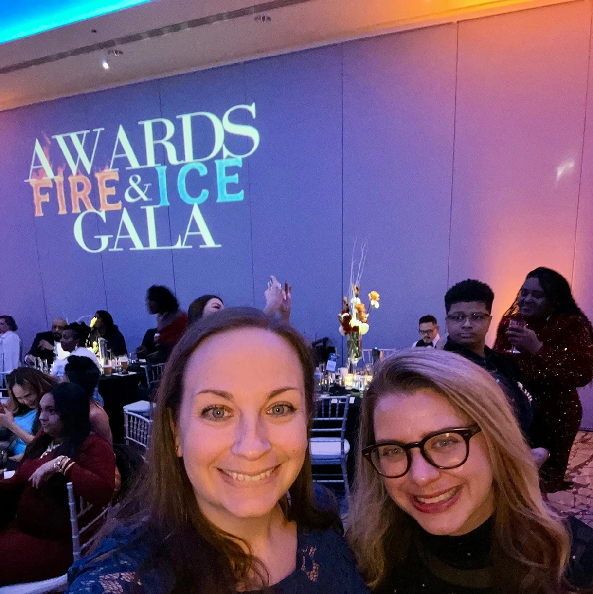 Two smiling women take a selfie in front of a wall projected with the text "Awards Fire & Ice Gala" at an indoor event.
