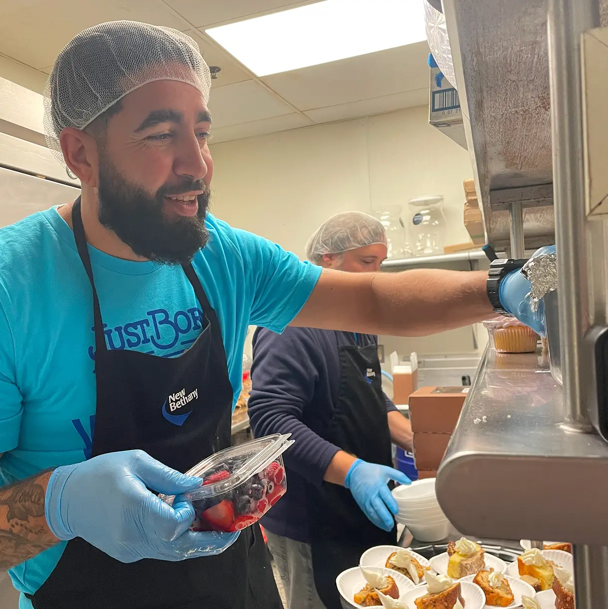A bearded male volunteer wearing a hairnet, a turquoise t-shirt, a black apron, and blue gloves smiles while holding a fresh container of mixed berries in a kitchen. 