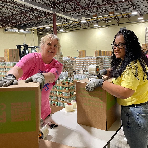Two female volunteers wearing work gloves smile as they assemble and seal cardboard boxes at a packing table in a large food bank warehouse.
