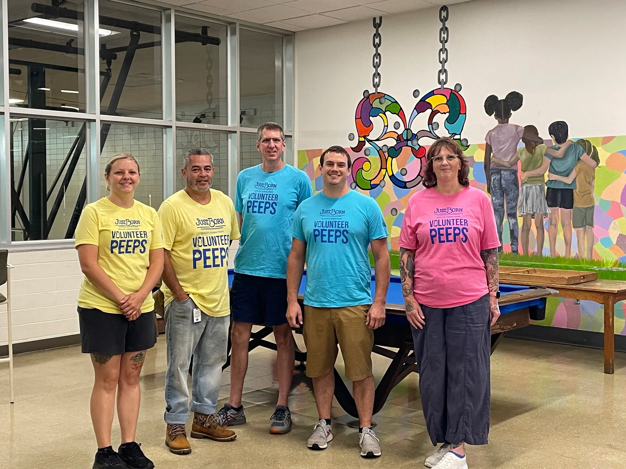Five volunteers wearing colorful ‘Volunteer Peeps’ shirts stand indoors in front of a colorful mural and game tables, posing together in a community space