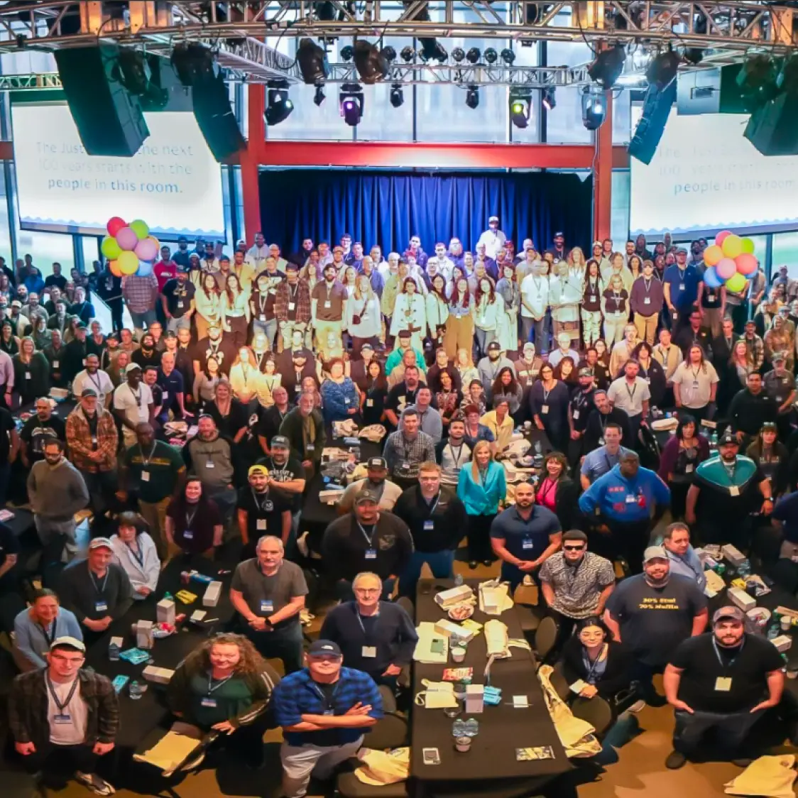 A group of people posing for a photograph at an indoor conference or event with a stage, lighting, and screens in the background.