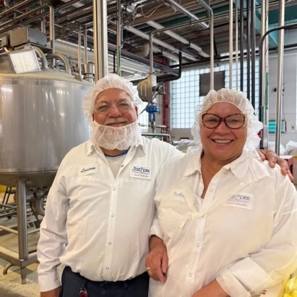 Two smiling workers in white lab coats and hairnets pose in front of industrial machinery at a Just Born factory.
