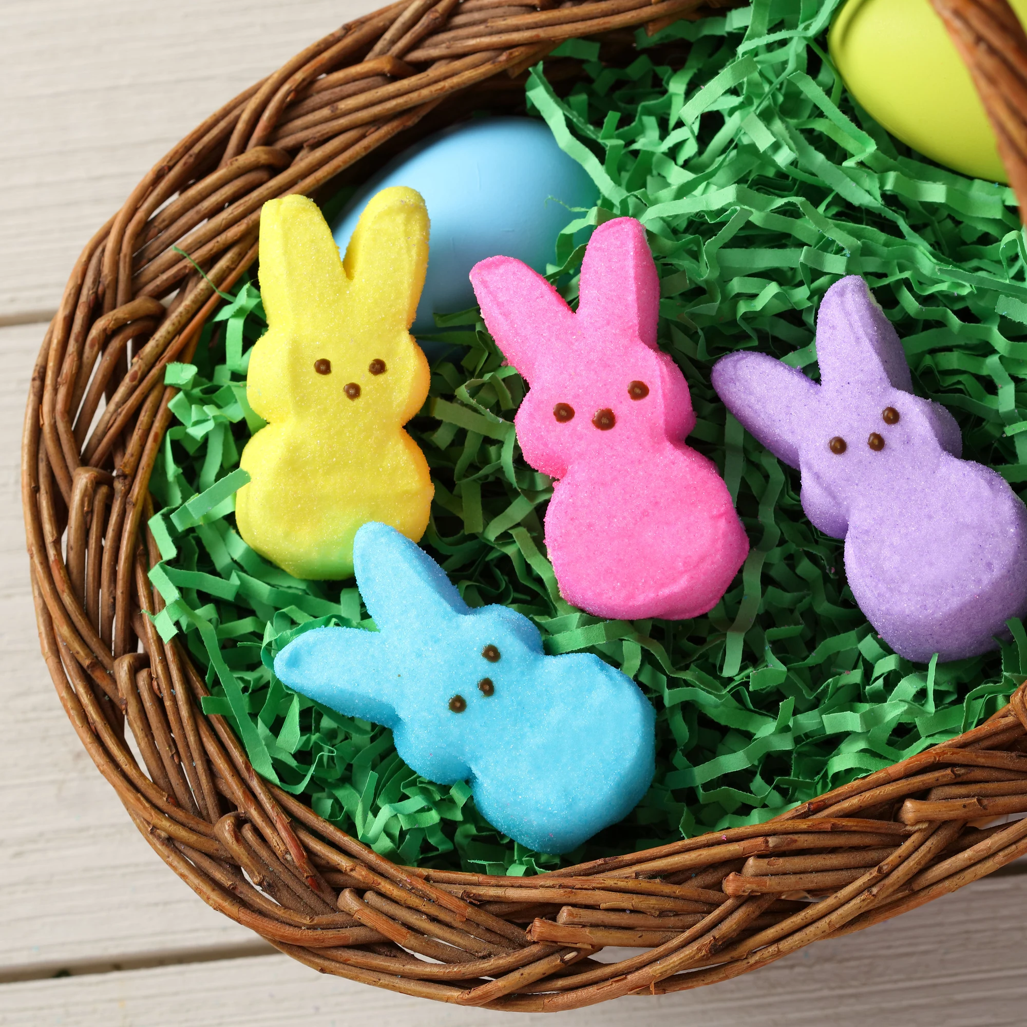 Wicker Easter basket with a gray plush bunny, Peeps marshmallow candies, and colorful eggs on green grass against a blue background.
